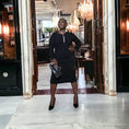 Load image into Gallery viewer, Plus-size woman wearing a black Boudicca Ruched Bodycon Midi Dress, styled with a sparkly necklace and black clutch, standing at the entrance of a restaurant.